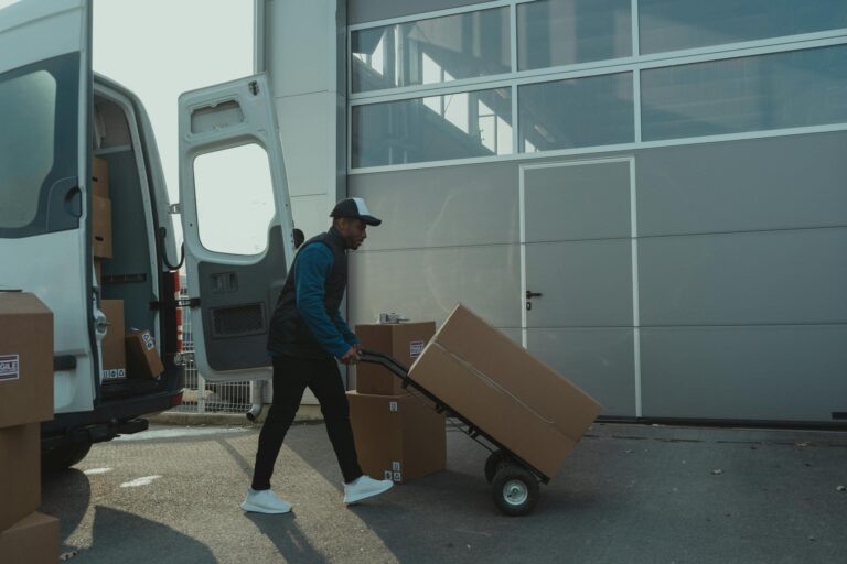 A delivery worker with a hand trolley managing a box shipment outside a warehouse.